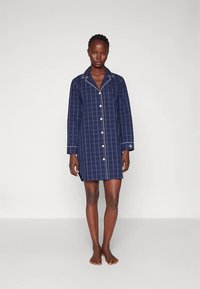 Mulher em pé descalça, vestindo uma camisa de noite de manga longa azul-marinho de xadrez, com detalhes em branco nas costuras.