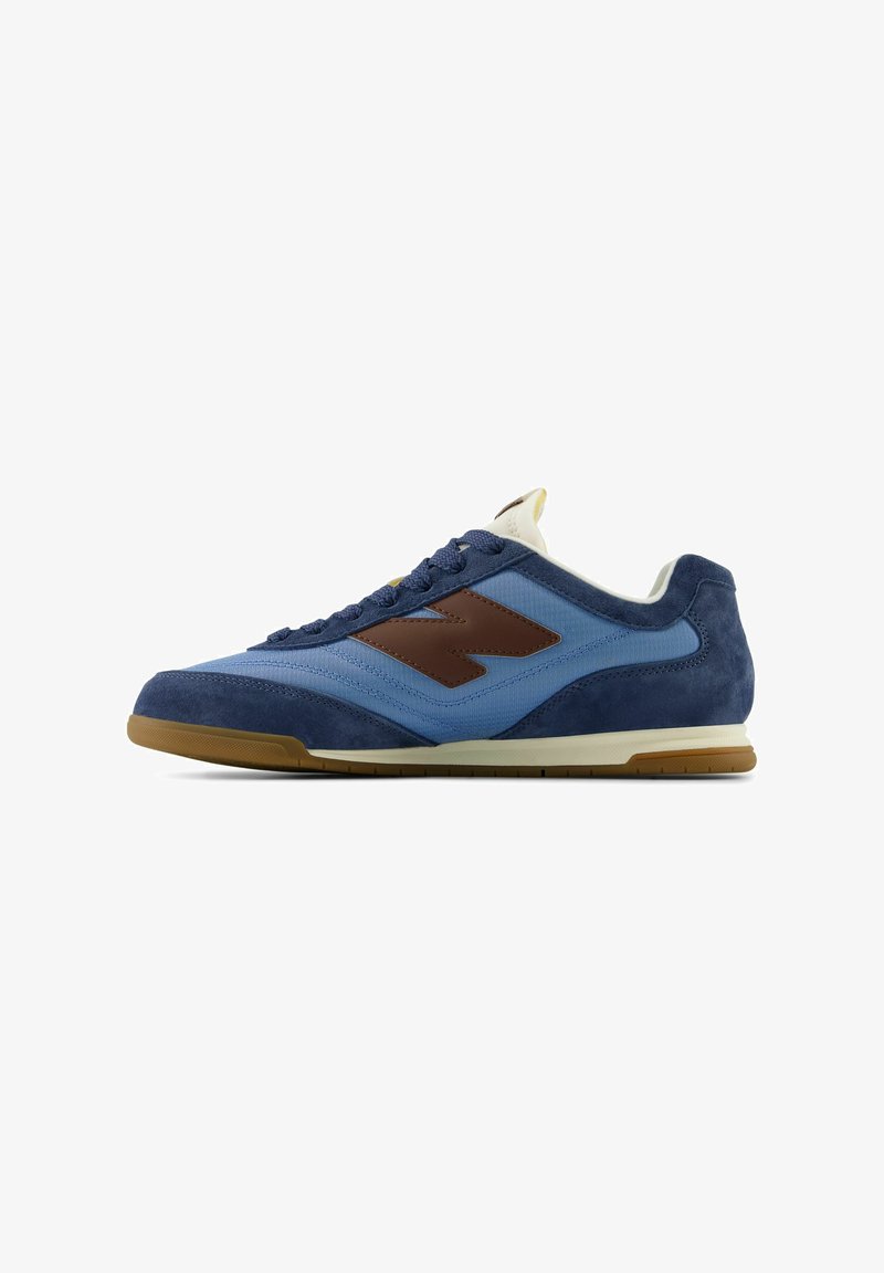 Low-top sneaker featuring a blue suede and fabric upper, dark brown leather accents, and a beige rubber sole. Lace-up design with a cushioned tongue.