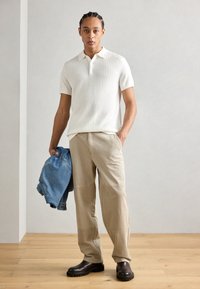 Witte geribbelde polo met korte mouwen, beige wijde broek en donkerbruine laarzen. Houdt een blauwe spijkerjack vast. Eenvoudig en modern ontwerp.