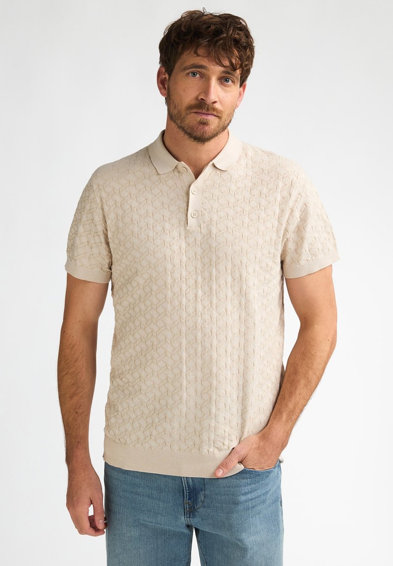 Polo beige en tricot avec motif géométrique texturé, manches courtes et col classique. Porté avec un jean bleu clair.