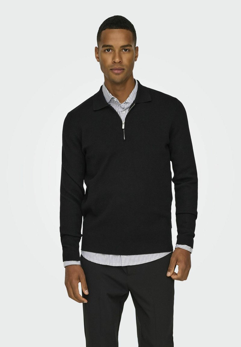 Schwarzer Pullover mit Reißverschlusskragen, langärmlig und mit taillierter Passform, getragen über einem weiß-schwarz gestreiften Hemd mit Kragen.