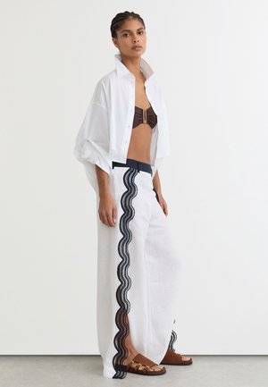 Kvinde iført hvide vidde bukser med sort blondekant, brun bandeau top, åben hvid skjorte og brune sandaler, stående mod hvid baggrund.