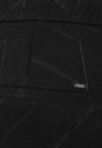 Una tela negra texturizada presenta patrones geométricos y un pequeño acento de logotipo metálico. El diseño incluye un bolsillo visible con costuras en contraste.