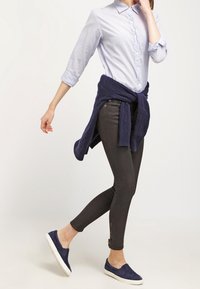 Camicia azzurro chiaro a righe con bottoni, jeans skinny scuri, maglione navy annodato in vita e scarpe slip-on navy con suola piatta.