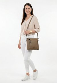 Lächelnde Frau mit beige Bluse, weißen Hosen, weißen Sneakers, goldener Uhr, die eine braune Umhängetasche über der linken Schulter trägt, vor weißem Hintergrund.