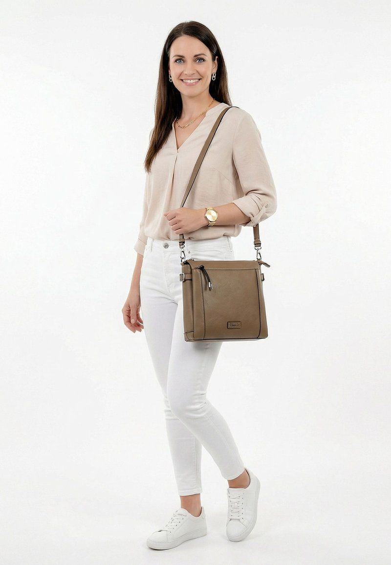 Lächelnde Frau mit beige Bluse, weißen Hosen, weißen Sneakers, goldener Uhr, die eine braune Umhängetasche über der linken Schulter trägt, vor weißem Hintergrund.