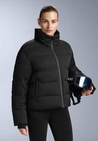 Crna puf jakna s visokim ovratnikom, zatvaranjem na zip i bočnim džepovima. Uparena s crnim tajicama. Ski naočale drži u ruci s reflektirajućim staklima.