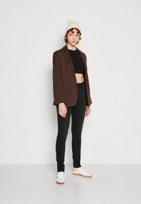 Smeđi sako preko crnog kratkog topa u kombinaciji s crnim skinny trapericama i bijelim tenisicama s narančastim detaljima. Model nosi bijelu kapu.