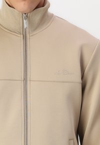 Beige Zip-Jacke mit hohem Kragen, aus glattem Stoff. Verfügt über subtile Nähte und ein geprägtes Logo auf der Brust.