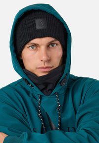 Man in een teal gevoerde jas met capuchon en een zwarte gebreide muts, armen gekruist, kijkt recht vooruit met een neutrale uitdrukking.