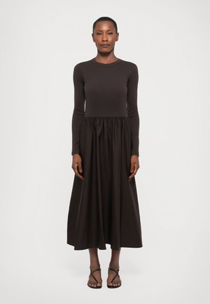 Long-sleeve brown dress made of soft fabric, featuring a fitted bodice and flowing skirt. Worn with simple black sandals.