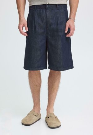 Denim shorts in dark blue, featuring front pleats, a button closure, and side pockets, paired with beige slip-on shoes.