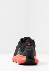 Chaussure de sport noire avec une partie supérieure texturée, présentant un accent rouge prononcé au talon et sur la semelle intermédiaire, avec une semelle épaisse et rembourrée.