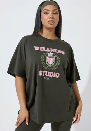 Femme aux longs cheveux foncés portant un T-shirt vert foncé ample avec l'inscription « Wellness Studio est. 1998 » ainsi qu'un bandeau et un pantalon assortis.