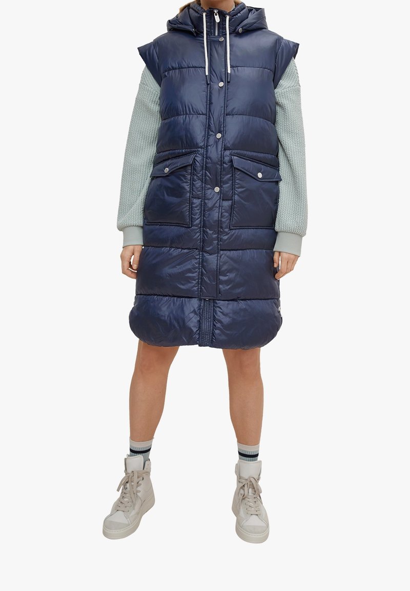 Marineblaue gesteppte Weste mit Kapuze, Druckknopfverschluss, Fronttaschen und gestepptem Design. Die Ärmel bestehen aus einem texturierten hellblauen Material.