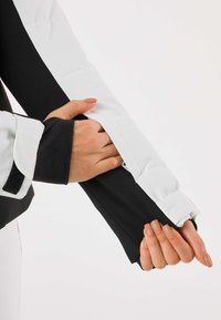 Sort og hvid atletisk jakke med en struktureret stof. Armen i fokus viser et tætsiddende design med velcrojusterbar manchet og en glat overflade.