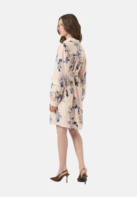 Abito rosa chiaro con stampa floreale blu, caratterizzato da maniche lunghe, vita con cintura e colletto, abbinato a scarpe con tacco alto marroni.