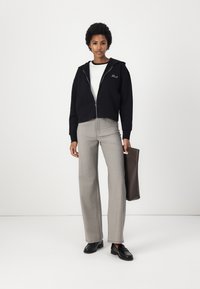 Schwarze, cropped Zip-Hoodie mit Kapuze, darunter ein weißes Crewneck-Shirt, graue, weit geschnittene karierte Hose, schwarze Loafers und eine braune Tragetasche.