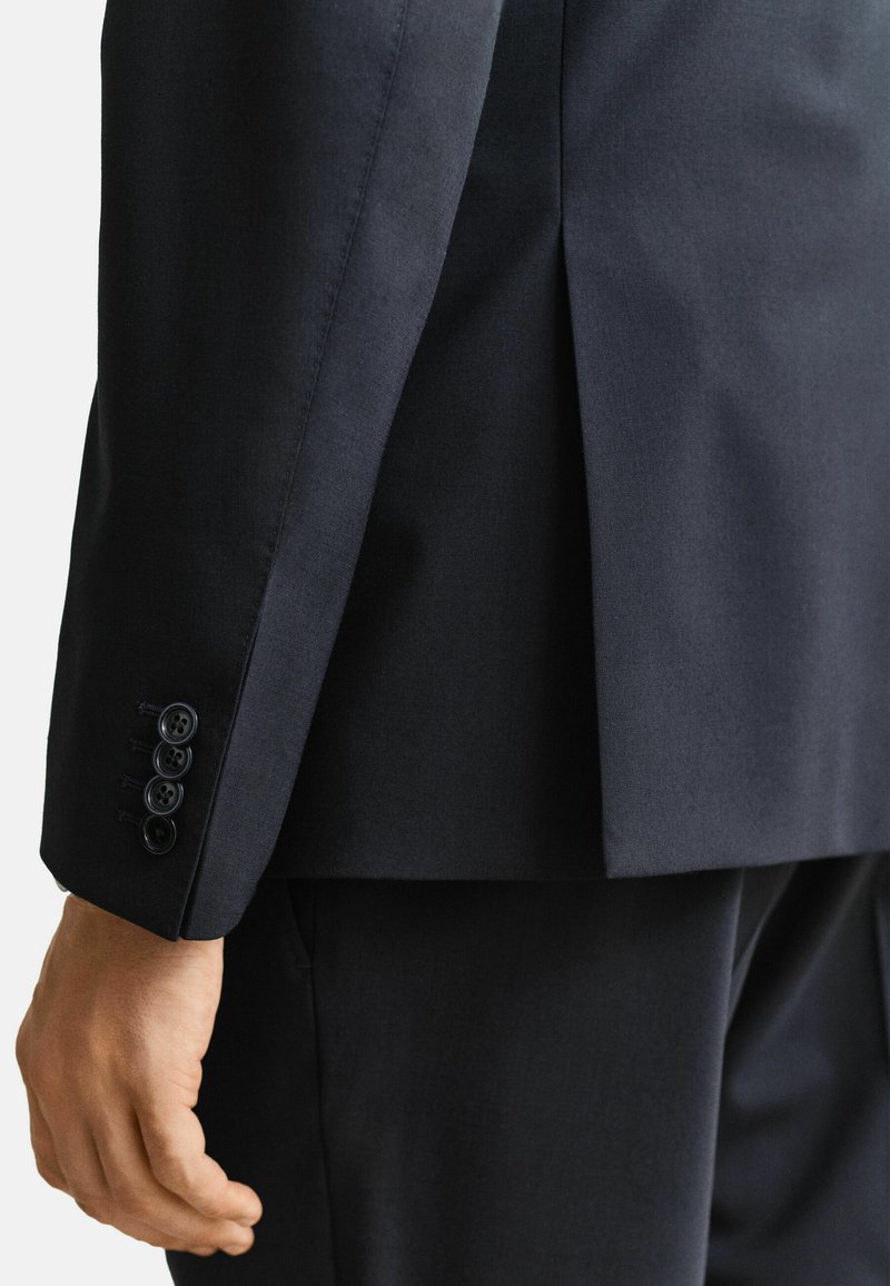 Bras gauche et main d'un homme portant une veste de costume bleu marine foncé avec quatre boutons sur le poignet, vus de dos.