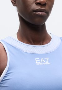 Κοντινή λήψη ενός ατόμου που φοράει ανοιχτό μπλε αμάνικη μπλούζα με λευκό λογότυπο "EA7 Emporio Armani" στο στήθος.