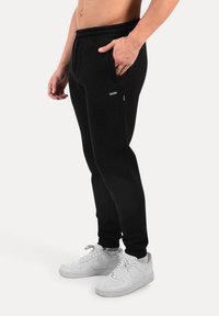 Muž v černých sportovních kalhotách s elastickým pasem a bočními kapsami, v kombinaci s bílými teniskami Nike a značkovými černými ponožkami.