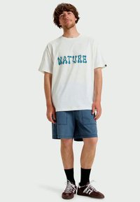 Λευκό βαμβακερό T-shirt με μπλε κείμενο "NATURE", σε συνδυασμό με μπλε βερμούδα και σκούρες κάλτσες. Καφέ αθλητικά παπούτσια με λευκές λεπτομέρειες.