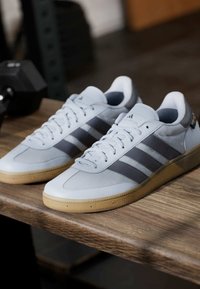 Lichtblauwe sportschoenen met drie donkergrijze strepen, een combinatie van mesh en gladde materialen, rubberen gumzool en platte veters, geplaatst op een houten oppervlak.