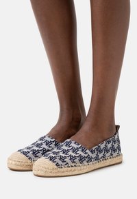 Närbild av ben som bär mörkblå och vita mönstrade espadrilloskor med vävda jute-sulor på en enkel bakgrund.