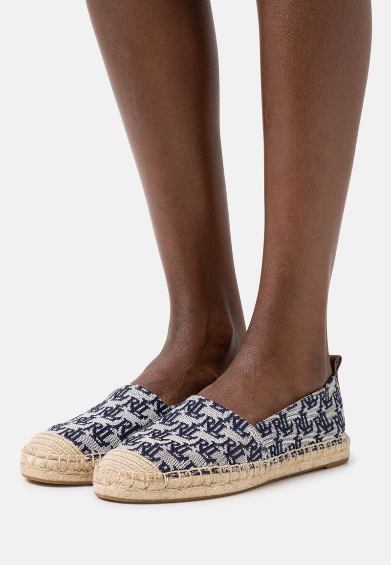 Närbild av ben som bär mörkblå och vita mönstrade espadrilloskor med vävda jute-sulor på en enkel bakgrund.