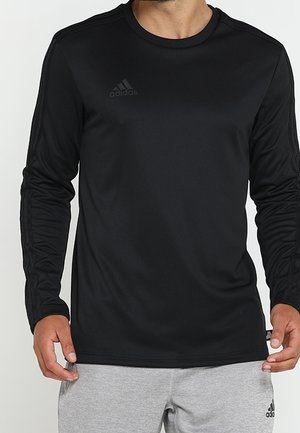 Mężczyzna w czarnej, długorękawowej koszulce Adidas i jasnoszarych spodniach dresowych Adidas, stojący z rękami swobodnie opuszczonymi wzdłuż ciała.