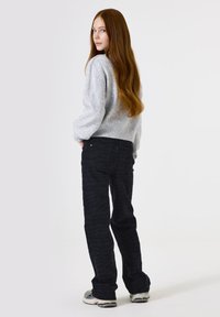 Hellgrauer Strickpullover und dunkel schwarze Jeans mit Zickzackmuster. Das Model steht seitlich mit langen Haaren und trägt weiße Sneakers.
