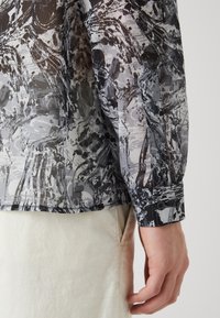 Camicia trasparente a righe nere e bianche con maniche lunghe e polsini abbottonati, abbinata a un tessuto di colore chiaro, che mostra diverse texture.