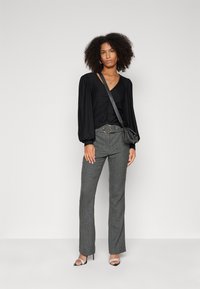 Black long-sleeve top with gathered front detailing, paired with gray textured trousers. Black handbag and silver heeled sandals complete the look.