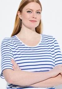 Bílý tričko s horizontálními modrými pruhy, kulatým výstřihem, krátkými rukávy a hladkou texturou. Modelka má dlouhé, rovné vlasy a nosí geometrické náušnice.