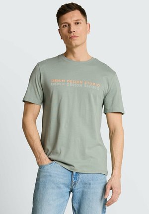 Mann trägt ein hellgrünes kurzärmeliges T-Shirt mit dem Schriftzug "DENIM DESIGN STUDIO" und hellblaue Jeans, steht mit einer Hand in der Hosentasche.