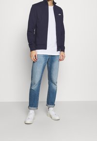 Uomo con giacca zip-up blu navy, t-shirt bianca, jeans chiari arrotolati, calzini bianchi e sneakers bianche in piedi contro uno sfondo semplice.