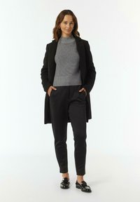Schwarzer langer Mantel über einem grauen gerippte Pullover, kombiniert mit schwarzen schmalen Hosen und glänzenden schwarzen Loafern, vor einem weißen Hintergrund.