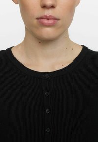Mustassa ribbattu neuletakissa on pyöreä kaula-aukko, ja edessä on kuusi mustaa nappia sekä pehmeä, tekstuuri kangas.