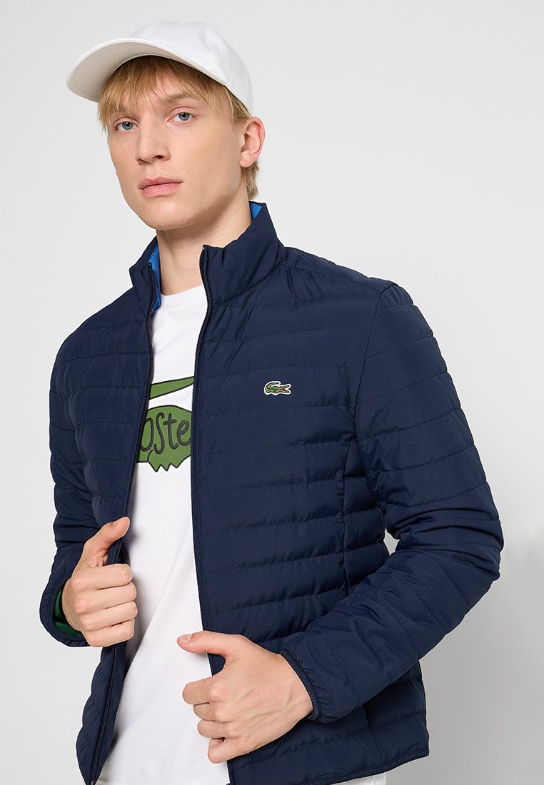 Veste matelassée bleu marine avec un col haut, fermeture à glissière sur le devant et logo brodé. Portée sur un t-shirt blanc avec des graphismes verts.