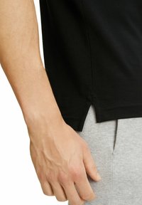 Czarna koszulka z krótkim rękawem z bocznymi rozcięciami, uszyta z miękkiego materiału, w połączeniu z jasnoszarymi spodniami dresowymi. Ręka spoczywa blisko szczegółu szwu.