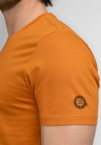 Camiseta de algodón naranja de manga corta con escote redondo y costuras decorativas. Un parche con logo circular es visible en la manga.
