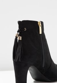 Černá semišová kotníková bota s vysokým podpatkem, s bočním zipem a detaily s třásněmi. Hladká textura a elegantní designové prvky.