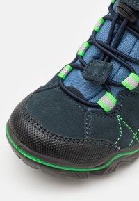 Chaussure en daim bleu foncé avec une semelle en caoutchouc texturé noir, des accents verts et argentés, des lacets élastiques et une languette rembourrée pour plus de confort.