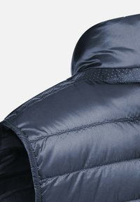 Gilet imbottito blu navy con una texture liscia e lucida e sezioni cucite. Presenta un colletto arrotondato e un accentuato a pois lungo la spalla.