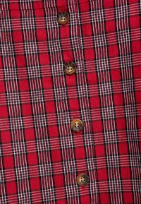 Il tessuto della camicia a quadri rossi presenta un motivo a griglia con linee nere e bianche; due bottoni marroni sono visibili sul fronte.