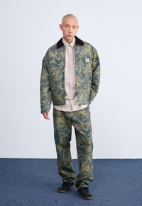 Giacca e pantaloni mimetici in tonalità di verde e giallo, con colletto nero, camicia beige con bottoni sotto, e scarpe scure.