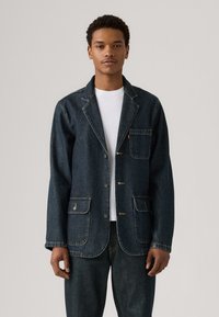 Σκούρο μπλε τζιν σακάκι με αυστηρό γιακά, τέσσερις μπροστινές τσέπες και κλείσιμο με κουμπιά. Διαθέτει ορατές ραφές και άνετη εφαρμογή.