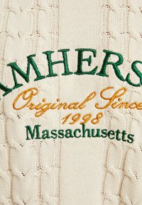 Krämfärgad kabelstickad tröja med grön och guld broderad text: "AMHERST," "Original sedan 1998," och "Massachusetts."