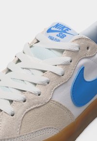 Άσπρο και μπεζ αθλητικό παπούτσι Nike SB Pogo Plus με μπλε σήμα, άσπρα κορδόνια και καφέ λαστιχένια σόλα, σε κοντινή όψη.