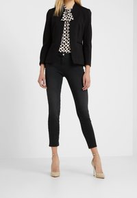 Blazer noir sur mesure porté sur un chemisier blanc à pois, associé à un jean skinny noir et des escarpins nude, présenté sur un fond neutre.